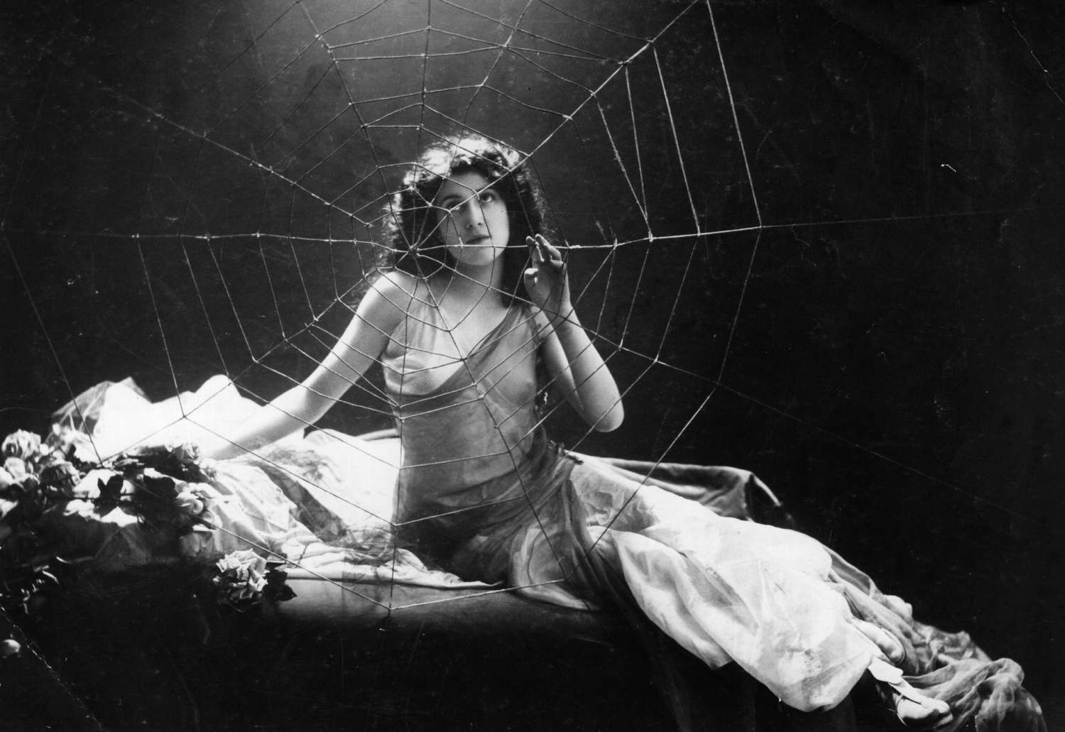 This black-and-white photograph portrays a woman seated on a surface draped with fabric and flowers. She is partially framed by a large spider web in front of her, which adds an ethereal, somewhat mysterious effect. The woman has wavy hair, wears a sheer dress, and gazes contemplatively through the web. The lighting is soft, casting shadows that enhance the dreamlike quality of the scene, evoking a sense of enigma or entrapment within the web. This image has a vintage, theatrical feel, possibly from the early 20th century.